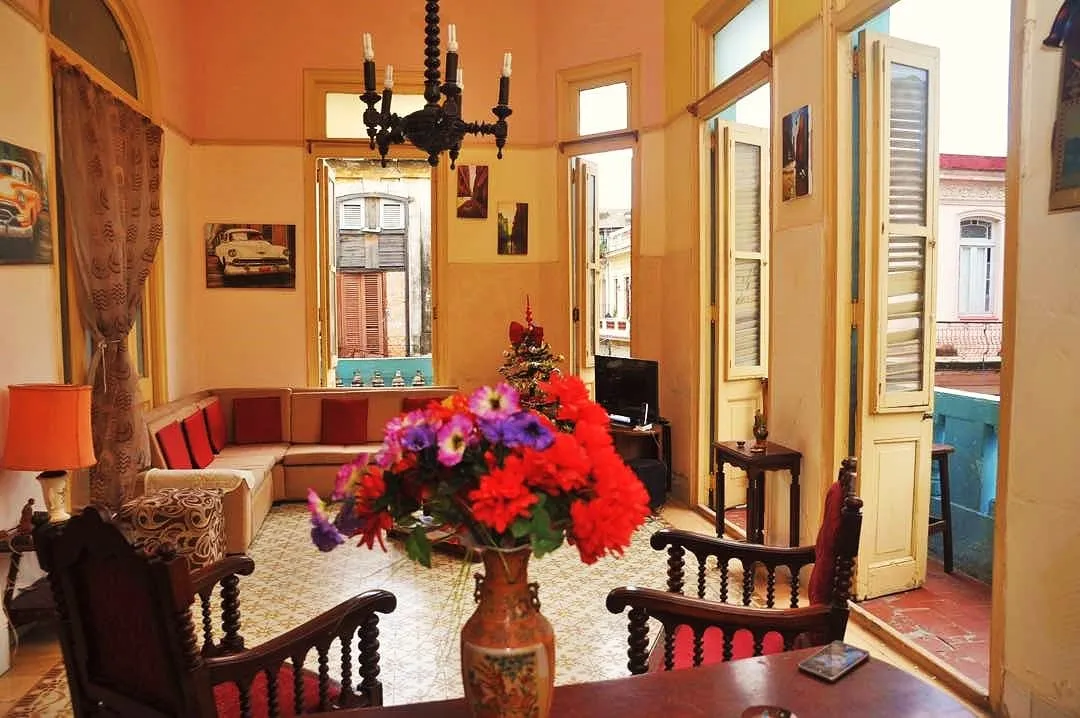 Centro Havana Large Colonial - Spacious colonial living room with high ceilings, ornate dark wood furniture, vibrant red velvet armchairs, colorful floral arrangements, and traditional patterned tile floors