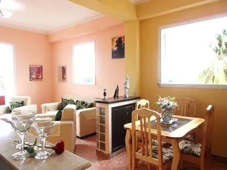 Miramar 1 Room - Bright open-plan living and dining area with cream furniture, green tropical accents, and palm trees visible through large windows